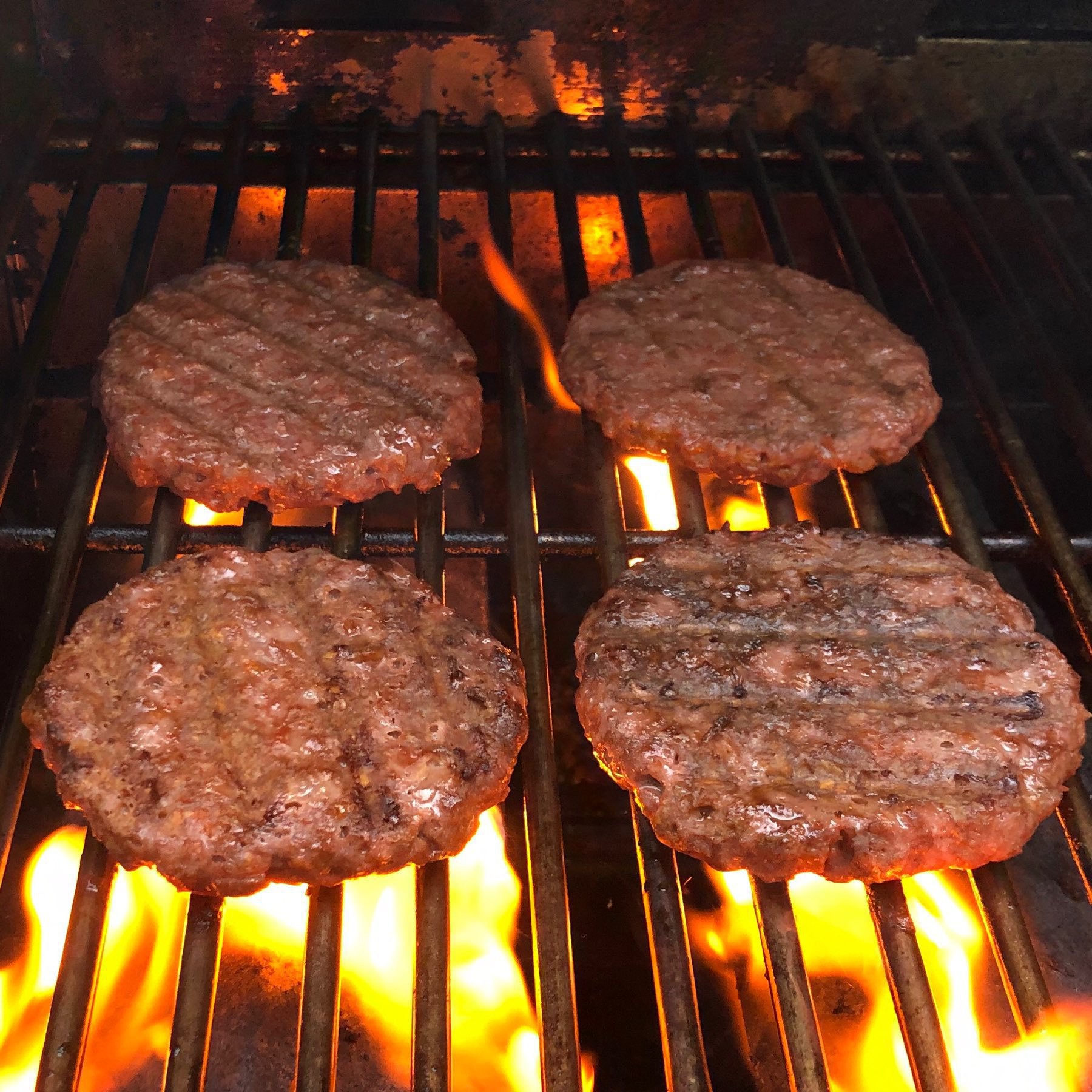 Four meat-like veggie burgers sizzling on a flame grill.
