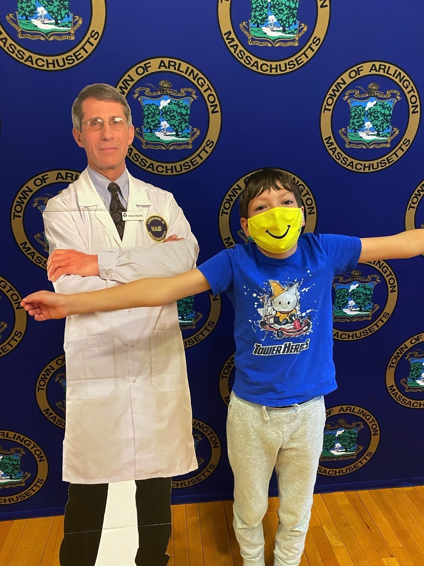 Cardboard cutout of Anthony Fauci alongside a 9yo boy with a happy face mask, arms reaching out.